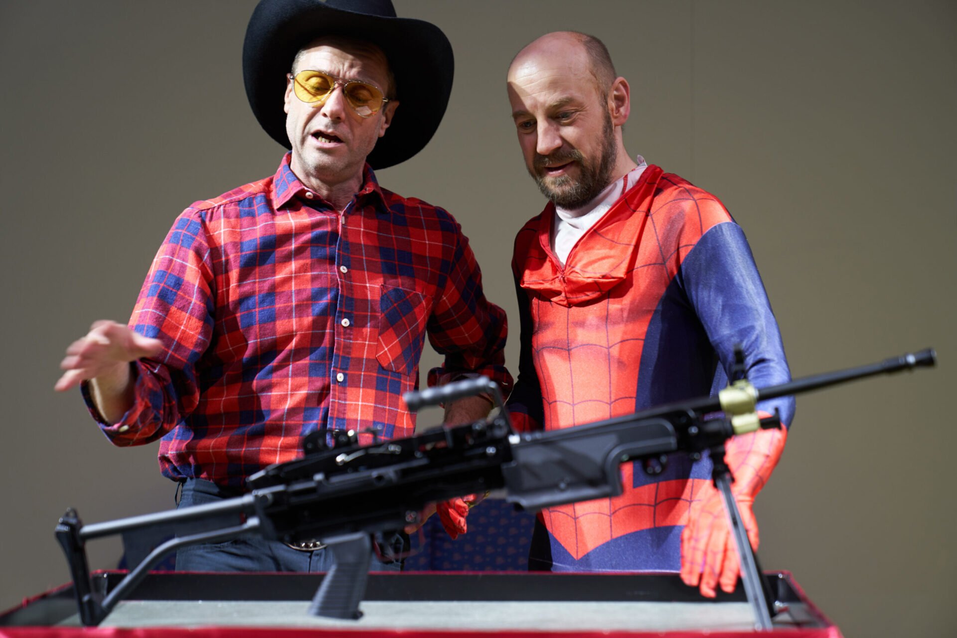 En mann med rutete skjorte og cowboyhatt og en mann i spidermanndrakt beundrer et automatvåpen