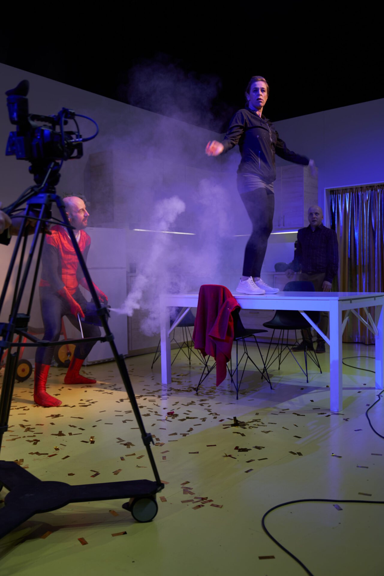 En kvinne danser på et bord mens en mann i spidermanndrakt står og ser på. En annen mann står i bakgrunnen.