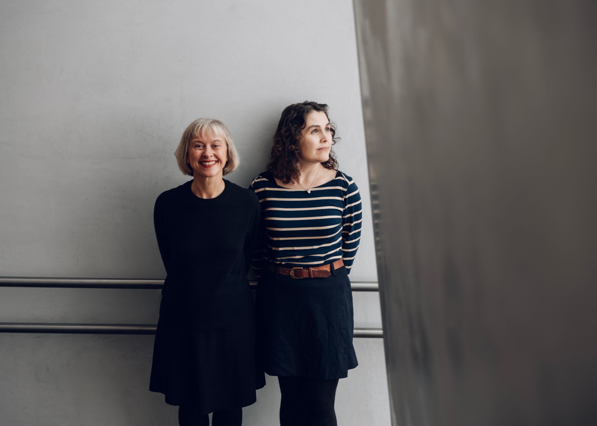 To kvinner står langs en grå vegg.