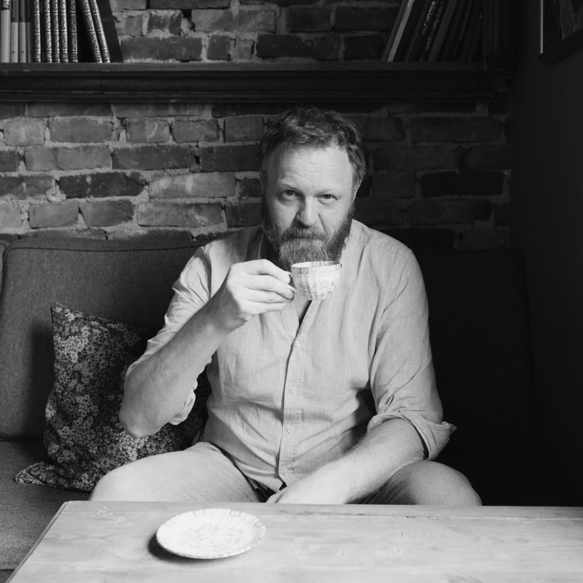 Sort-hvitt bilde av mann med skjegg og lys skjorte som sitter og drikker kaffe ved et trebord.