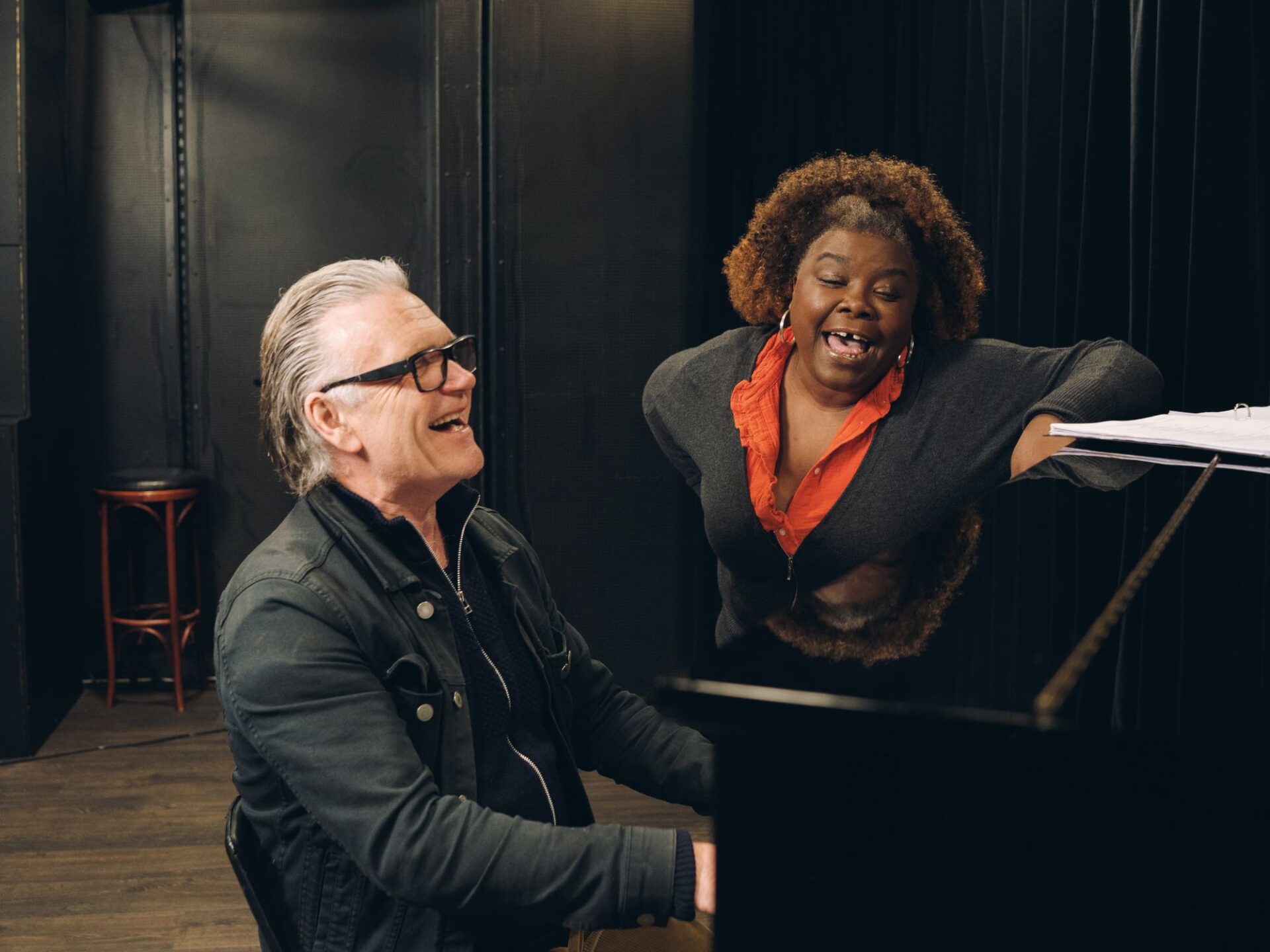 En mann med mørk jakke og mørk zip-up sitter og spiller ved et piano. En kvinne i mørk cardigan og oransje bluse står ved pianoet og synger.