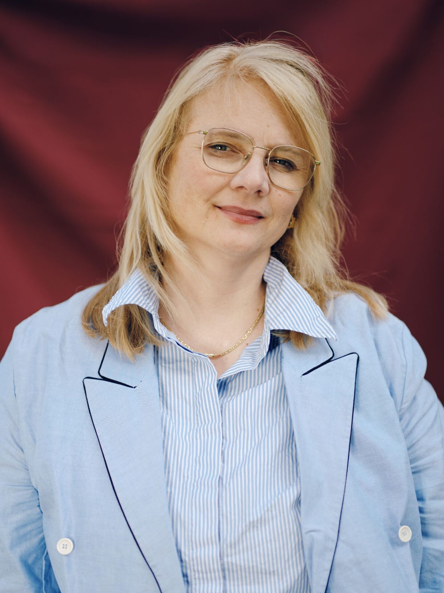 En kvinne med blondt hår og lyseblå skjorte og jakke smiler mot kamera.